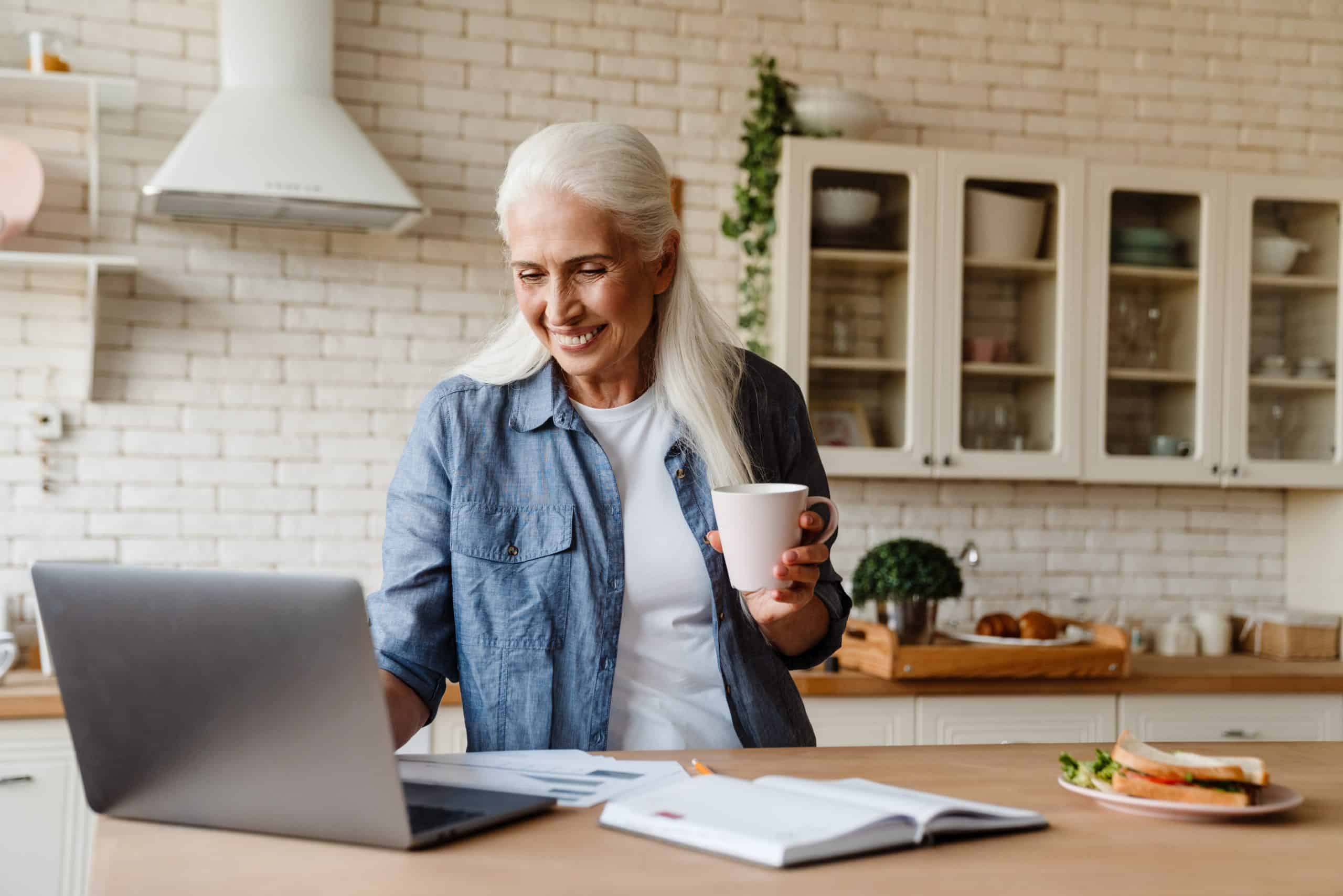 Senior smiling woman working on her laptop in the kitchen, holding cup of tea